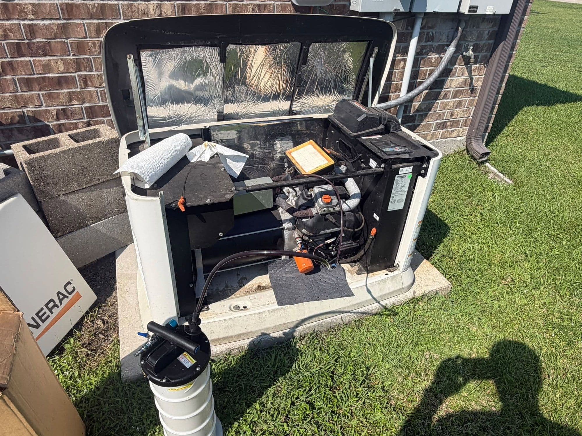 A Generac standby home generator opened for service with an oil extractor connected, on a concrete pad beside a brick house