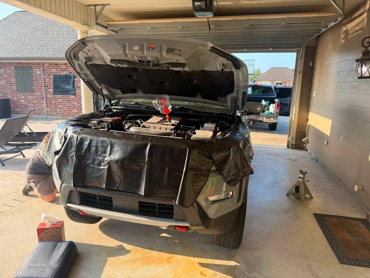 A dark SUV with the hood up in a carport, with a fender cover protecting the paint